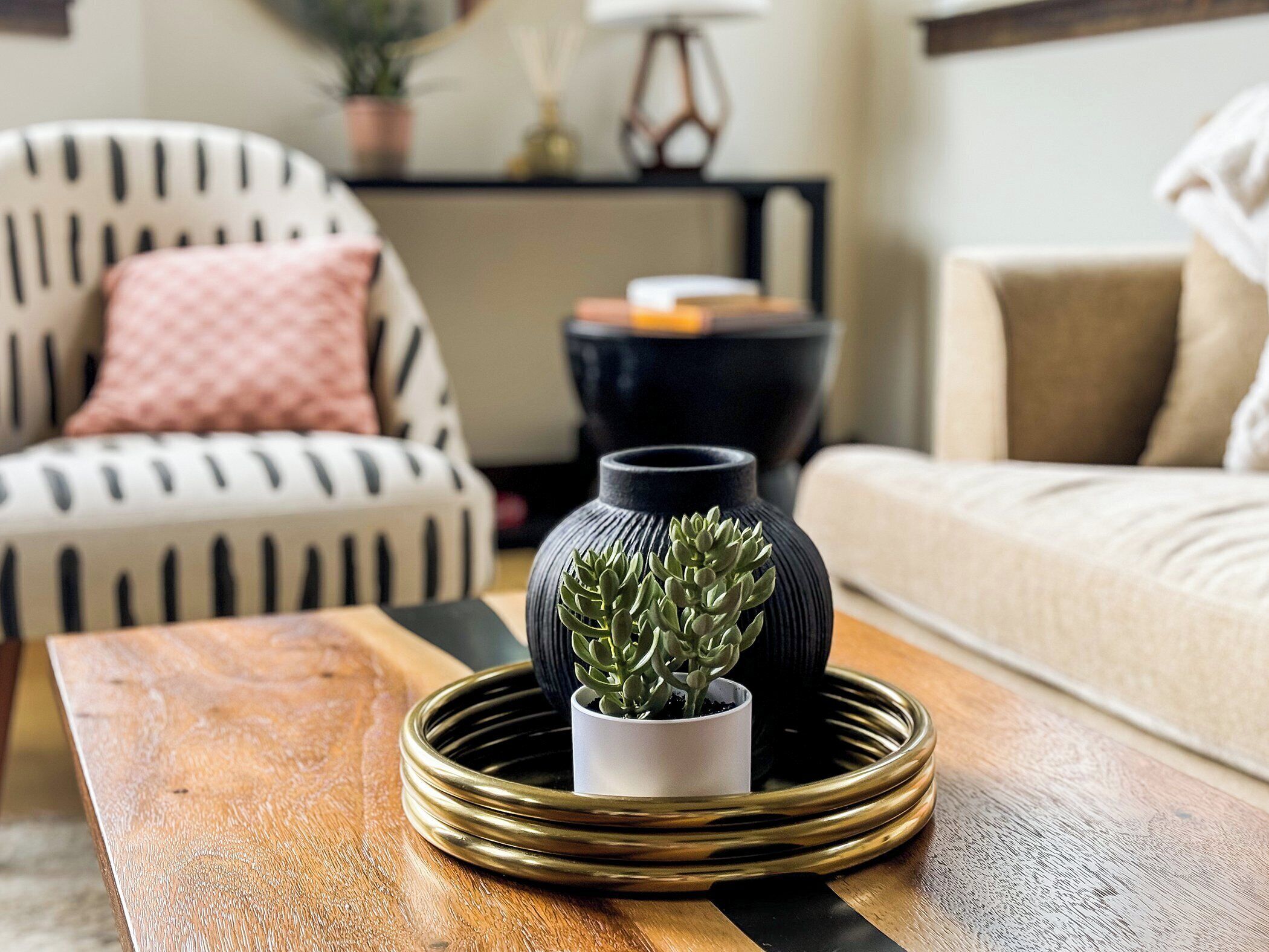 Uma sala de estar com uma mesa de centro de madeira, um vaso preto com uma planta suculenta, uma poltrona listrada e um sofá bege.