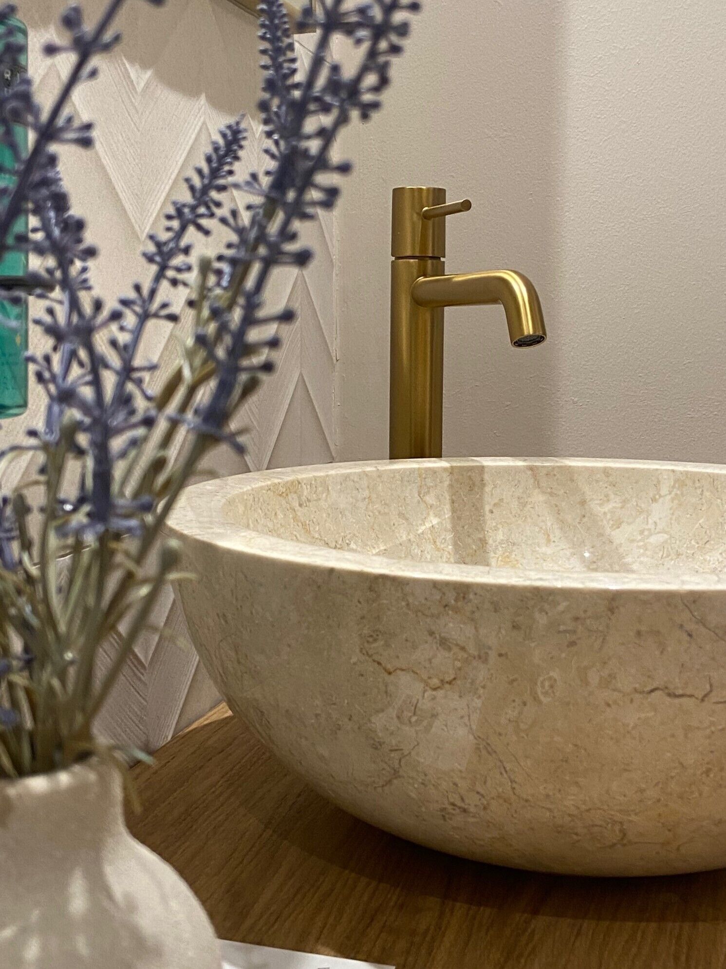 A round, beige ceramic sink with a gold faucet, set against a white wall with a decorative pattern.