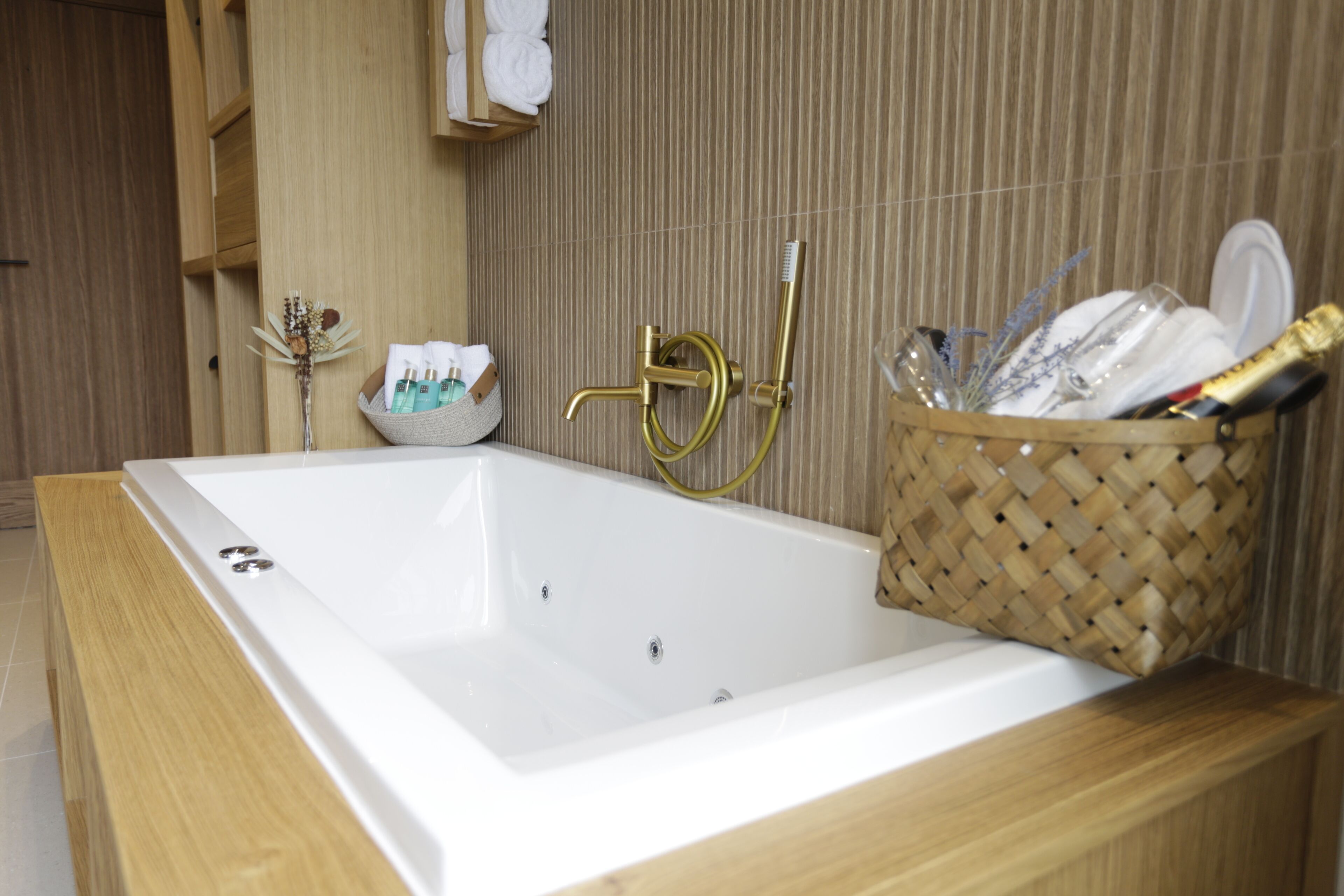 A modern bathroom with a large white bathtub, a gold faucet, and a wicker basket filled with toiletries.