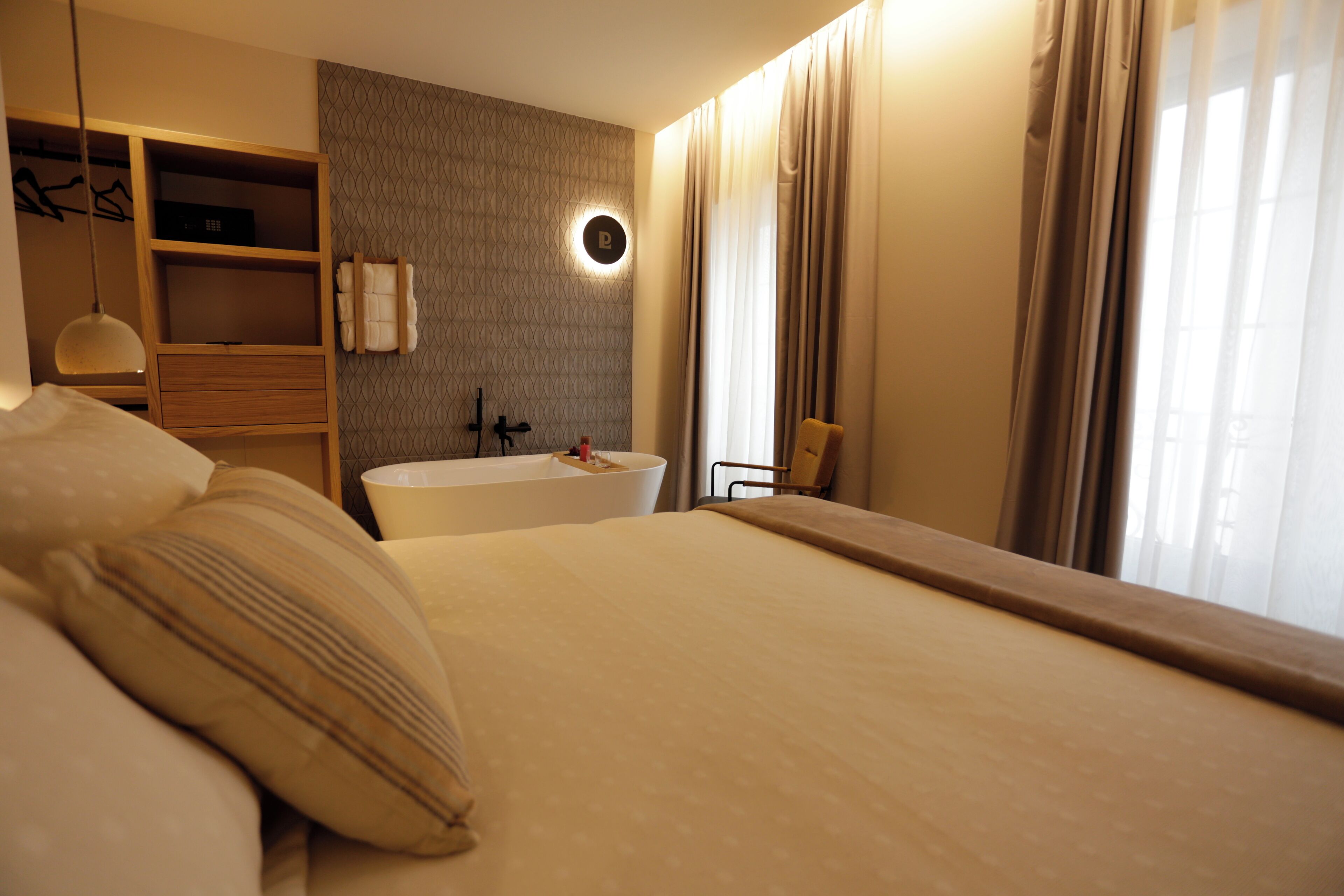 A hotel room with a large bed, a built-in bathtub, a wooden wardrobe, and a round wall-mounted light fixture.