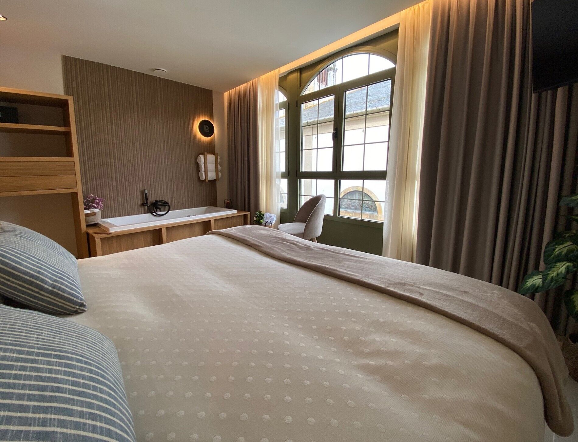 Een hotelkamer met een groot bed, een nachtkastje met een lamp, een raam met gordijnen en uitzicht op buiten.