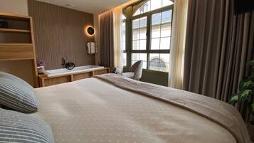 A hotel room with a large bed, a bedside table with a lamp, a window with curtains, and a view of the outside.