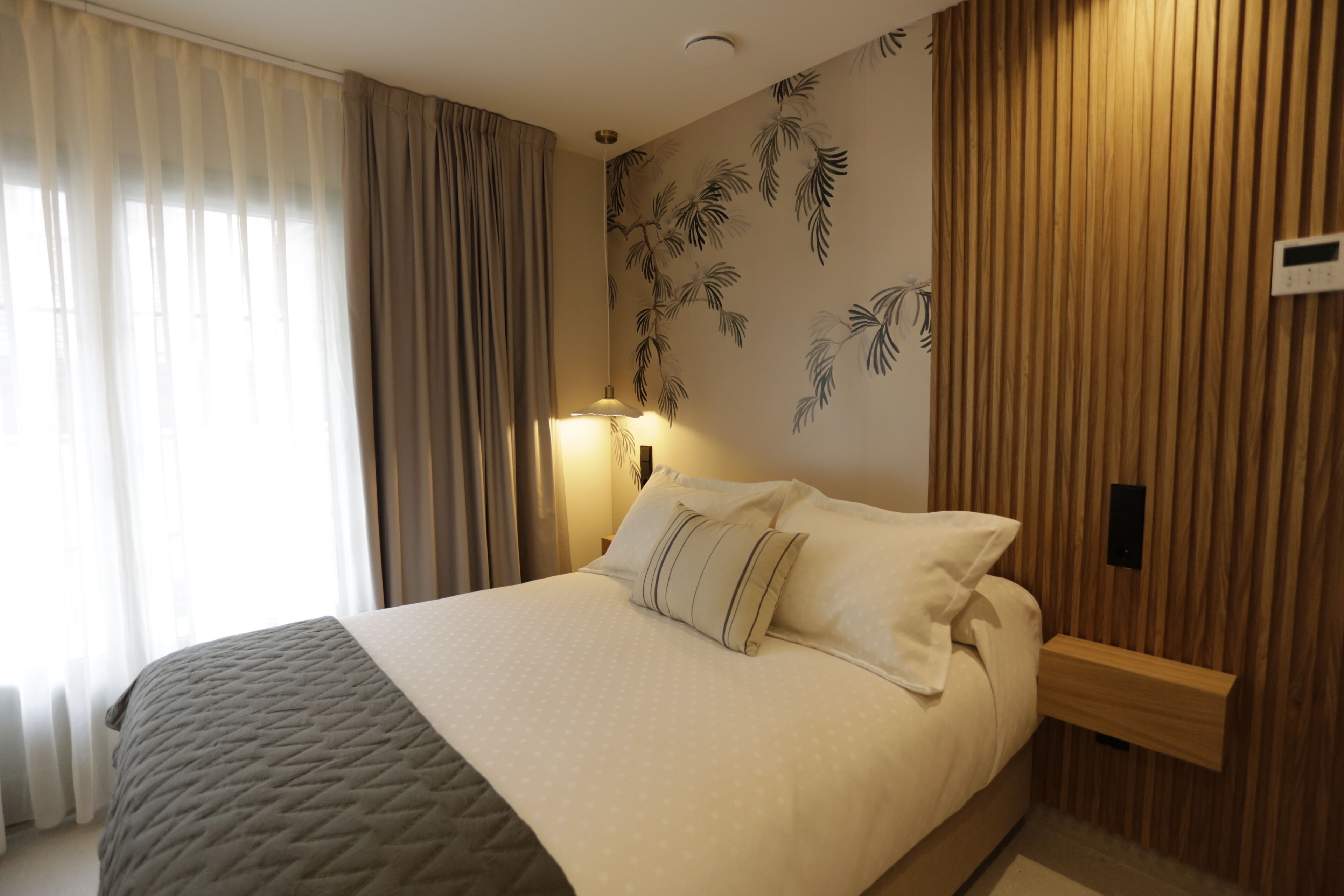 A neatly made bed with a quilted bedspread and pillows, a wall-mounted nightstand with a lamp, and a wooden panel wall with a palm leaf pattern.