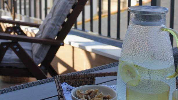 A wicker table with a glass pitcher, a bowl of nuts, and a green napkin. In the background, there is a white building with a bell tower and a view of a hill. - Hotel Puerto de Luarca  (Valdes)