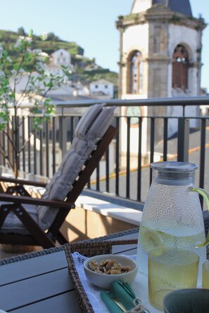 A wicker table with a glass pitcher, a bowl of nuts, and a green napkin. In the background, there is a white building with a bell tower and a view of a hill. - Hotel Puerto de Luarca  (Valdes)