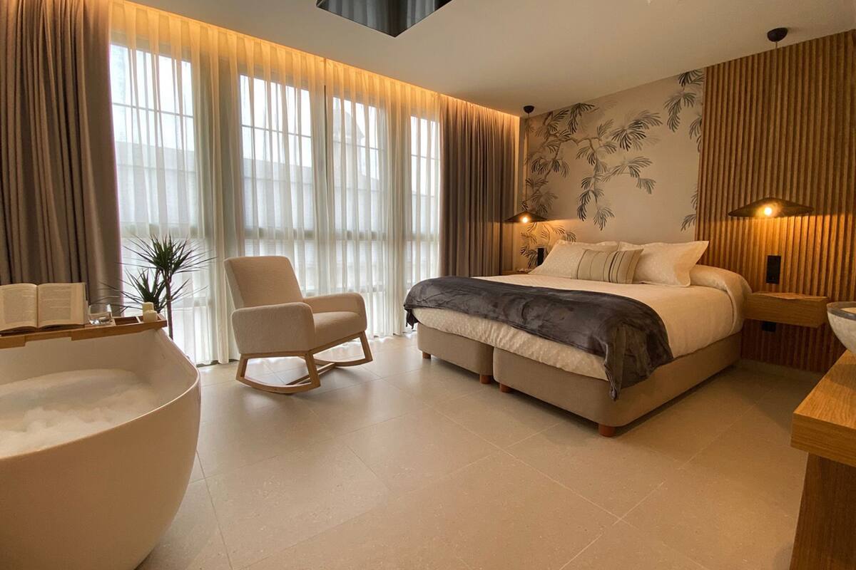 Ein modernes Schlafzimmer mit einem großen Bett, einem Schaukelstuhl, einer Badewanne und einem großen Fenster mit transparenten Vorhängen.