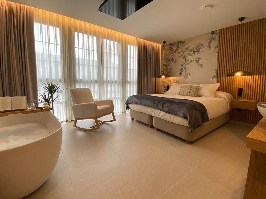 Un dormitorio moderno con una cama grande, una mecedora, una bañera y un ventanal con cortinas translúcidas.