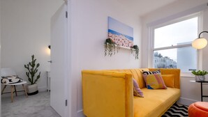 Un salon moderne avec un canapé jaune, un tableau représentant une scène de plage et une fenêtre donnant sur la ville.