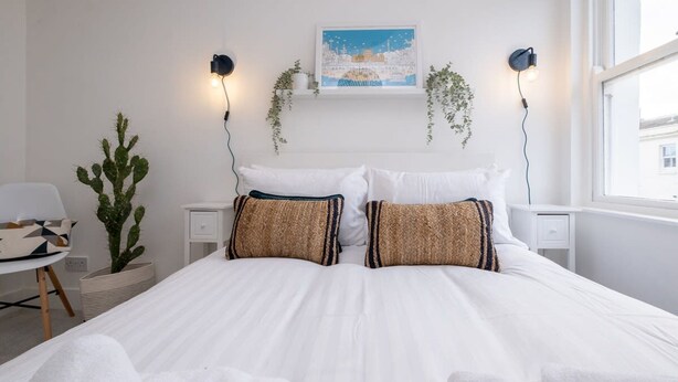 Un lit bien fait, avec du linge de lit blanc et deux coussins décoratifs, un cactus et un luminaire fixé au mur.