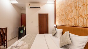 A hotel room with a bed, a wooden headboard, a small desk with a refrigerator, and a door. - The Lodgers Hotel and Banquet Sector 38 (Gurugram)