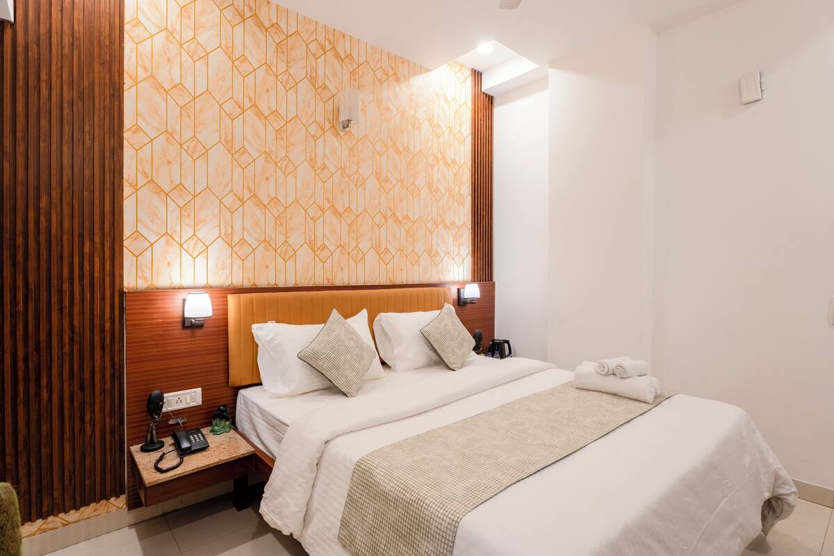 Una habitació d'hotel amb un capçal de llit de fusta, una paret amb un disseny de motius geomètrics, un llit amb roba de llit blanca, una taula de nit amb una làmpada, un telèfon i una petita planta.