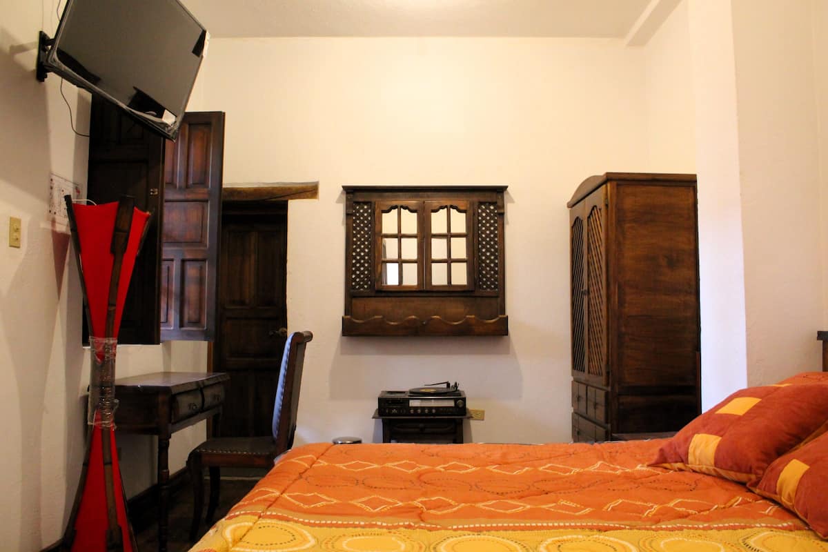 A bedroom with a bed, a wooden wardrobe, a small table with a radio, and a TV mounted on the wall.