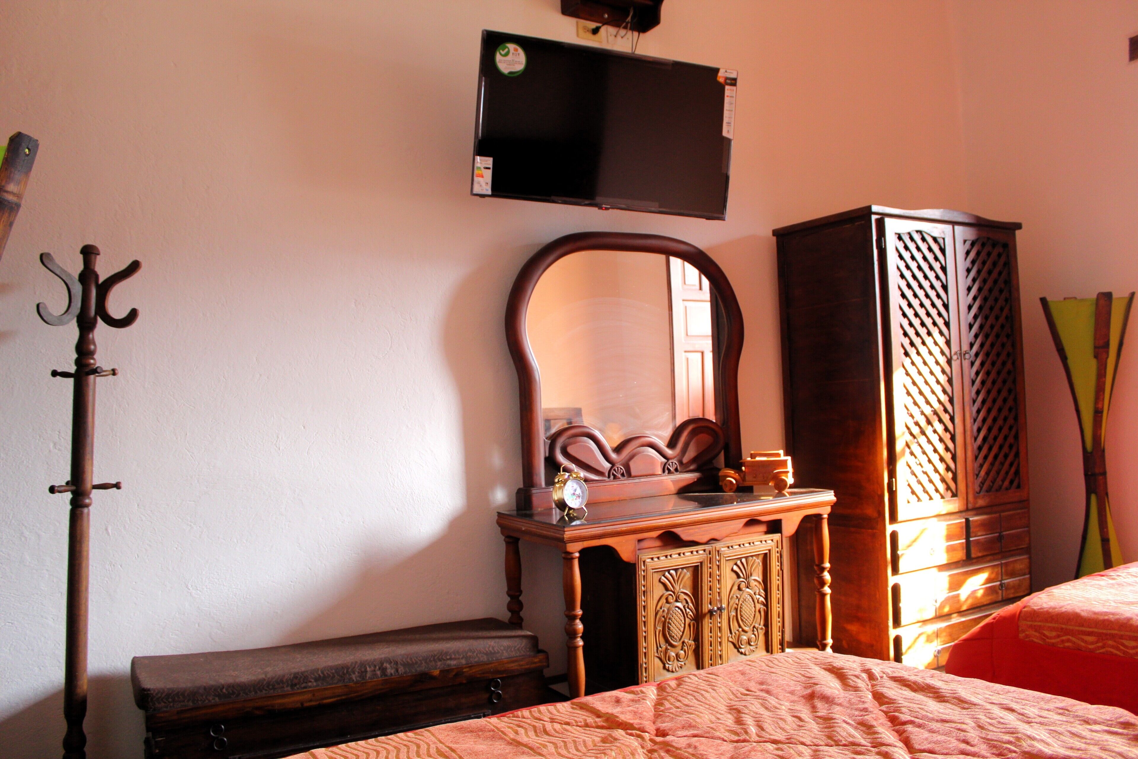 Une chambre à coucher avec un lit, une commode en bois, un miroir, un porte-manteau et un téléviseur fixé au mur.