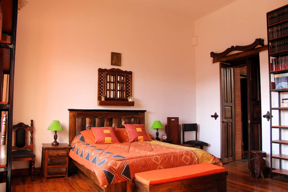 A traditional wooden bed with a headboard, two bedside tables with lamps, a bench at the foot of the bed, and a wooden door with a bookshelf to the right.