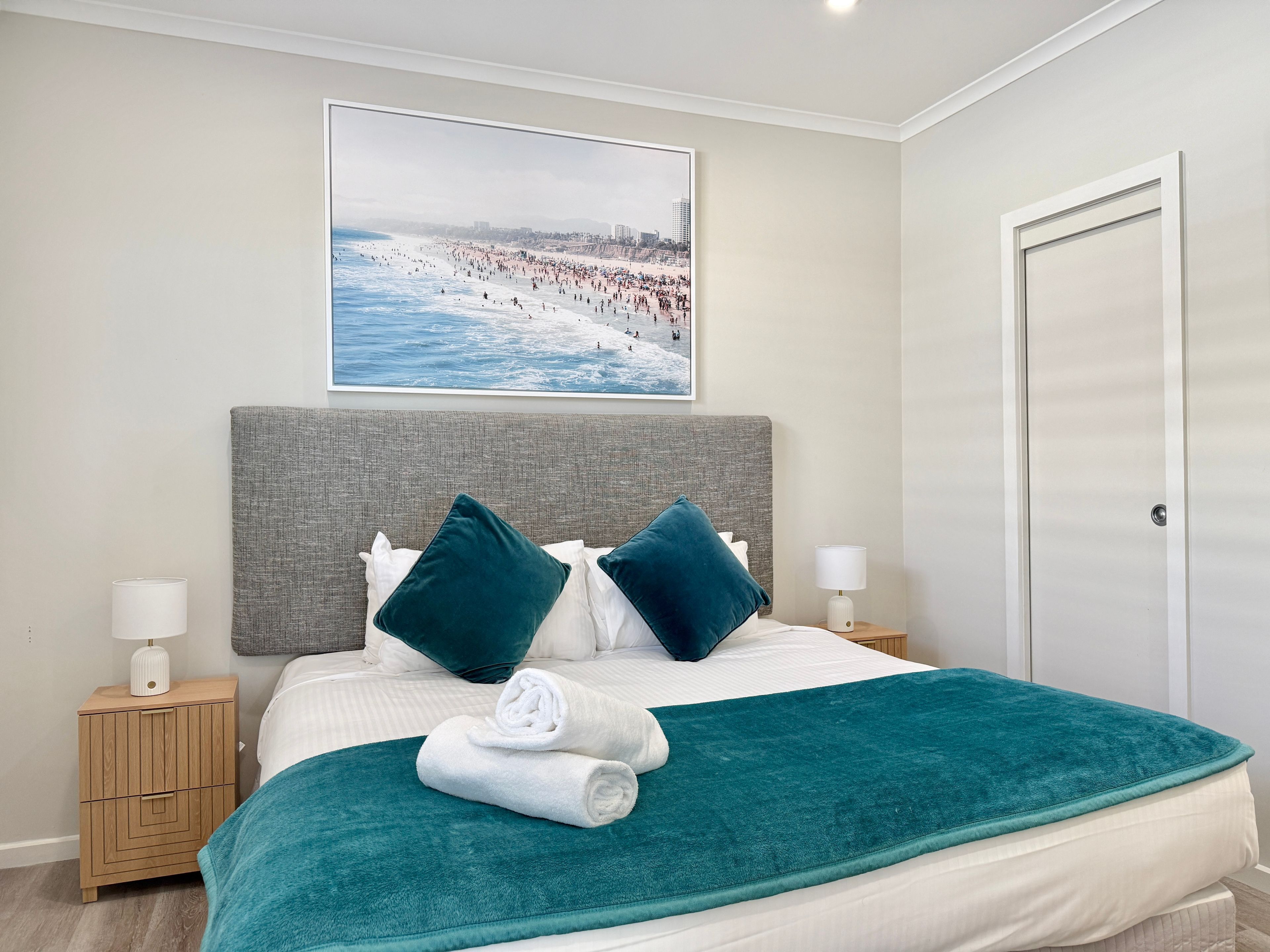 Une chambre d’hôtel avec un lit, une table de chevet, une lampe, un tableau représentant une scène de plage et une porte.