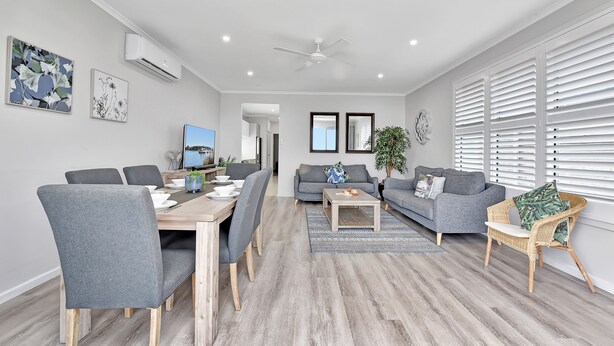 A modern living room with a dining area, grey sofas, a coffee table, and a TV. - Ocean Mist at Caves Beach Villas (Caves Beach)