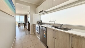 A modern kitchen with a central island, stainless steel appliances, and a view to the dining area. - Ocean Mist at Caves Beach Villas (Caves Beach)