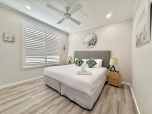A bedroom with a bed, a ceiling fan, a window with blinds, a bedside table with a lamp, and a wall-mounted artwork.