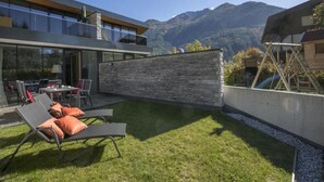 Ein modernes Haus mit einer Steinwand, einer Terrasse mit Liegestühlen und Blick auf die Berge.