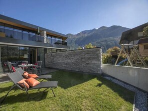 Een modern huis met een stenen muur, een terras met lounge stoelen en uitzicht op de bergen.