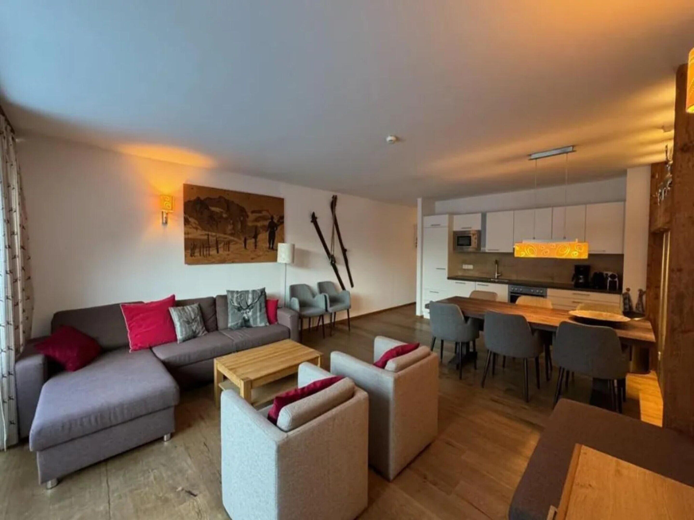 Ein modernes Wohnzimmer mit einer Couch, Hocker und Sesseln, ein Essbereich mit Tisch und Stühlen sowie eine Küche mit Schränken und Geräten.