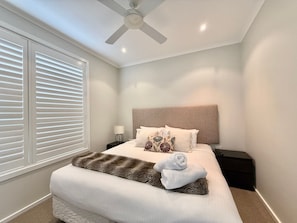 Un dormitorio con cama, mesita de noche, lámpara, ventilador de techo y ventana con persianas.