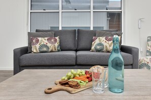 Un salon avec un canapé gris, des coussins à motifs, une table basse en bois sur laquelle sont posés des collations et des boissons, et une fenêtre avec des stores.