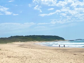 A beach with a sandy shore, a grassy hill in the background, and a clear sky.