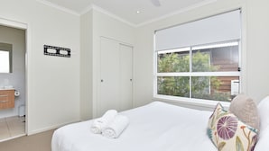 A hotel room with a bed, a window with a view of greenery, a bathroom with a toilet and sink, and a ceiling fan. - Beach Escape at Caves Beach Villas (Caves Beach)