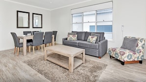 A modern living room with a grey sofa, a dining table with chairs, a coffee table, and a large window. - Beach Escape at Caves Beach Villas (Caves Beach)