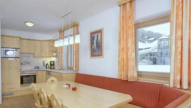 Uma sala de jantar com mesa e cadeiras de madeira, uma cozinha com eletrodomésticos básicos e uma janela com cortinas.