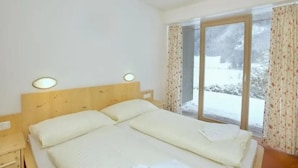 Quarto com cabeceira de madeira, cama com roupa de cama branca, mesa de cabeceira e vista para uma paisagem nevada através de uma janela com cortinas florais.