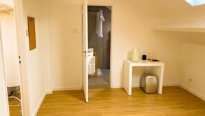 Una habitación con suelo de madera, una puerta blanca, una pequeña mesa blanca con un electrodoméstico plateado y vistas a un baño con ducha.