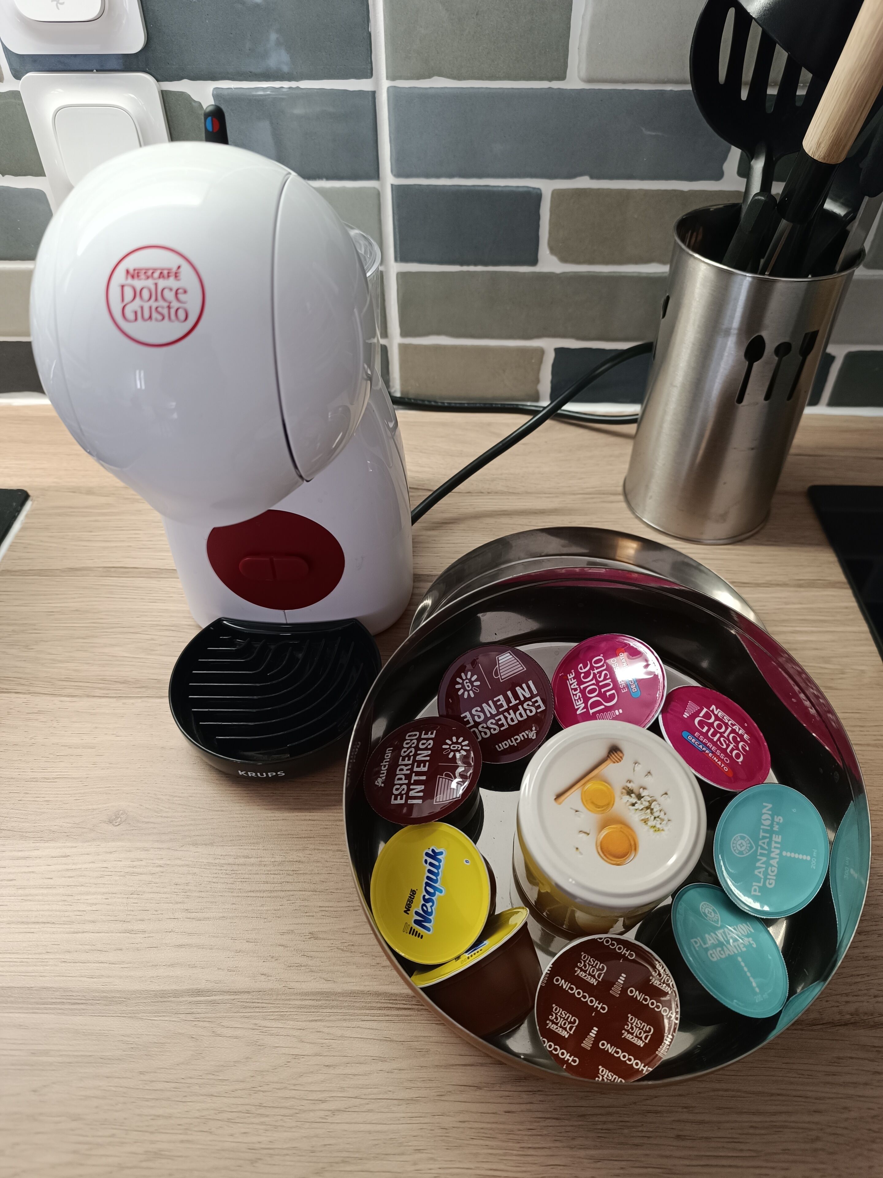 Eine Küchenarbeitsplatte mit einer Nescafé Dolce Gusto Kaffeemaschine, einem Edelstahlbehälter voller verschiedener Kaffeekapseln und einem Metall-Utensilienhalter.
