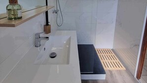 Une salle de bain moderne avec un lavabo blanc, une douche équipée d’un pommeau de douche à main et une étagère en bois sur laquelle est posée une bouteille.