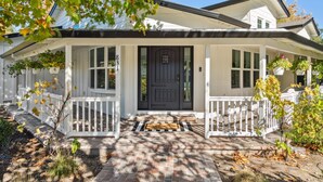 A two-story house with a dark front door, white railings, and a covered porch with hanging planters.