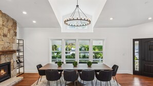 A dining area with a stone fireplace, a chandelier, and a view of greenery through large windows.