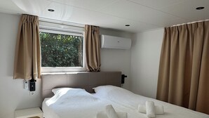 A bedroom with a large bed, a window with curtains, an air conditioning unit, and a small bedside table. - 3bedroom, heated pool, 20min to beach, kidsproof (Heinkenszand)