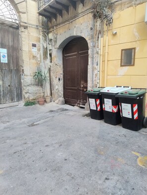 A street with a closed brown door, a yellow wall, and trash bins labeled 'VETRO' and 'CARTA & CARTONE'.