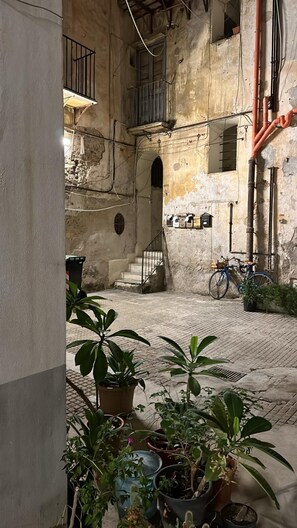 An outdoor courtyard with a cobblestone floor, potted plants, a bicycle, and a staircase leading to an upper level.