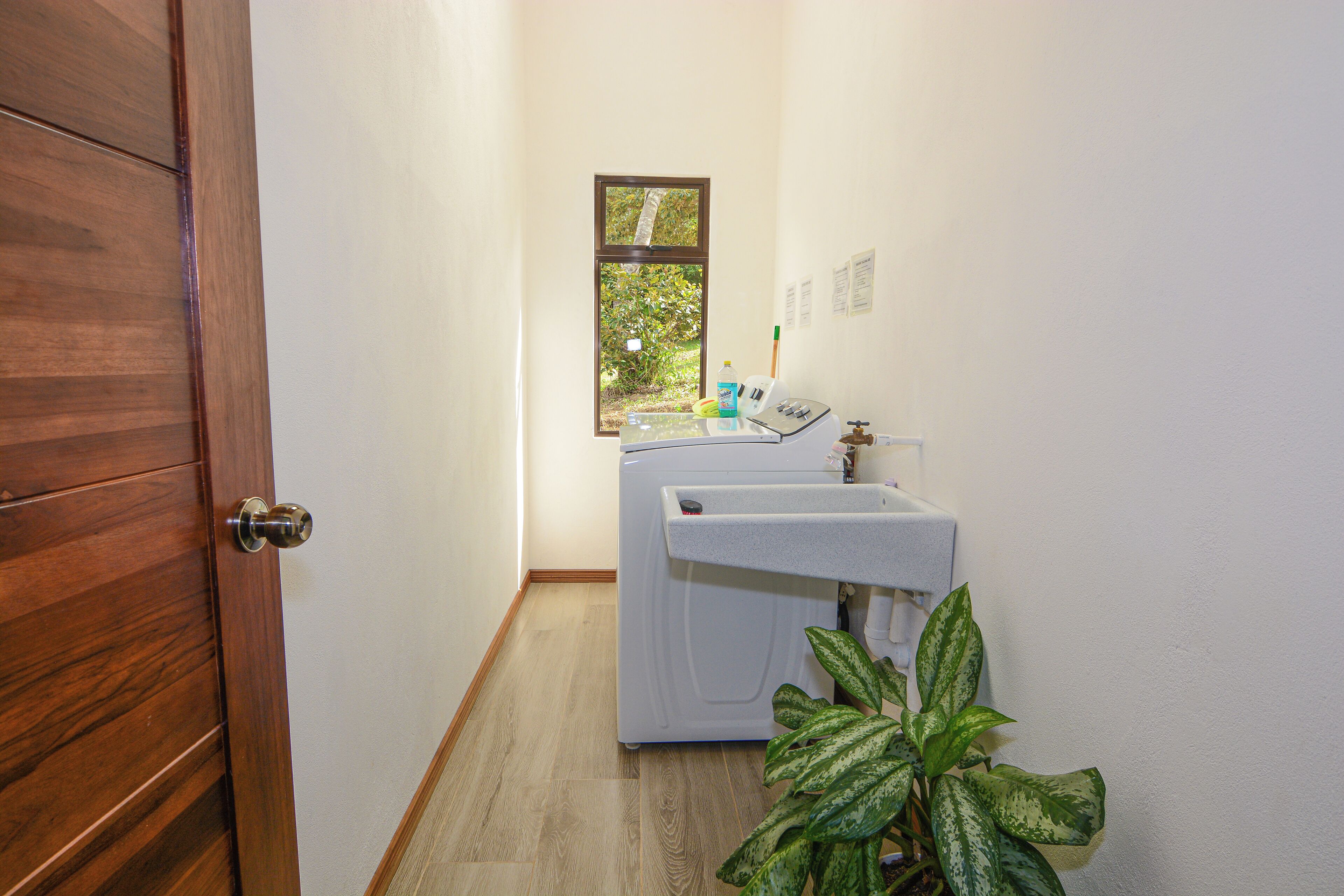 A laundry area with a washing machine and dryer, a window with a view of greenery, and a wooden door.