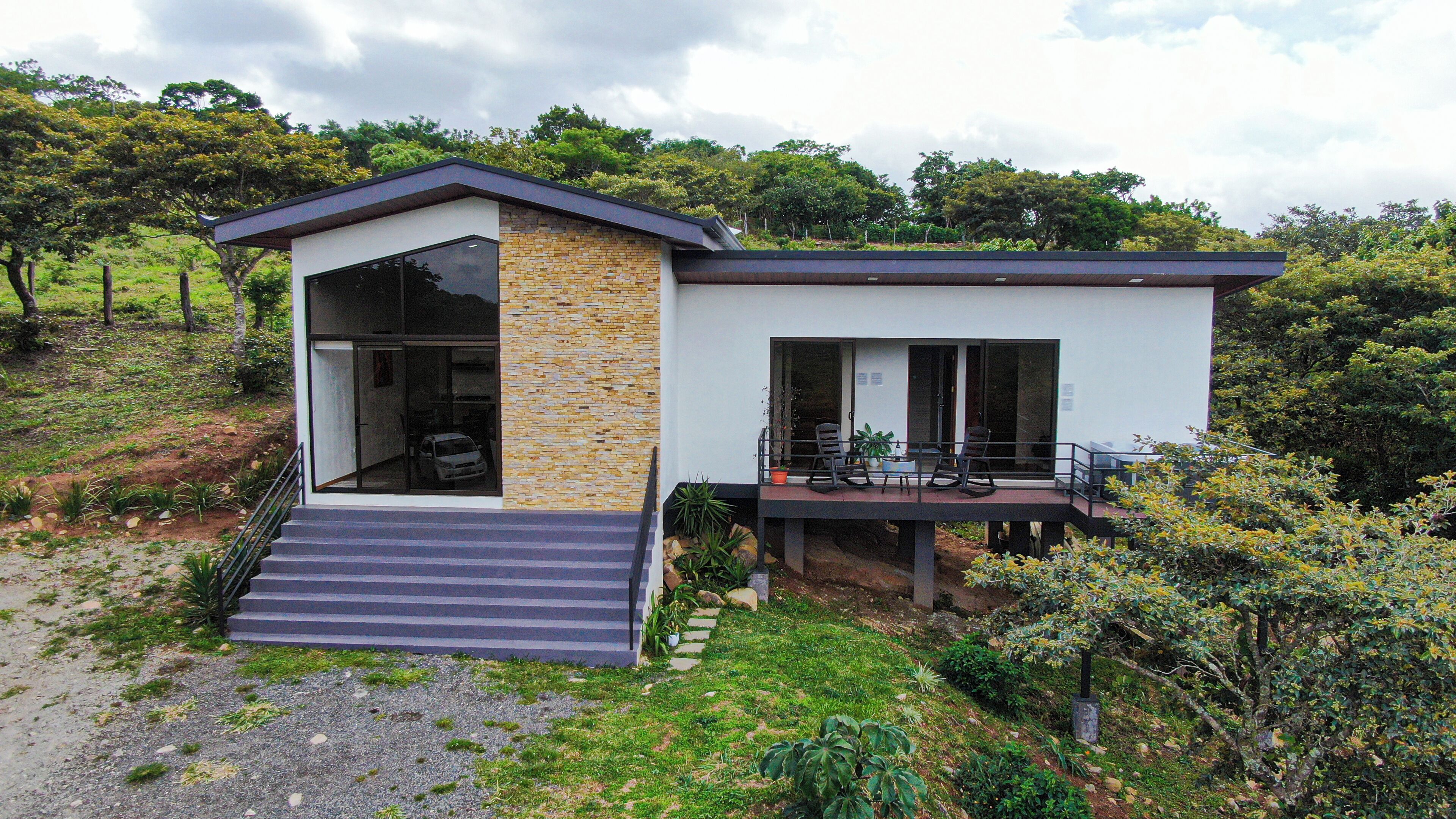 A modern house with a large glass door entrance, a small porch with outdoor seating, and a stone wall.