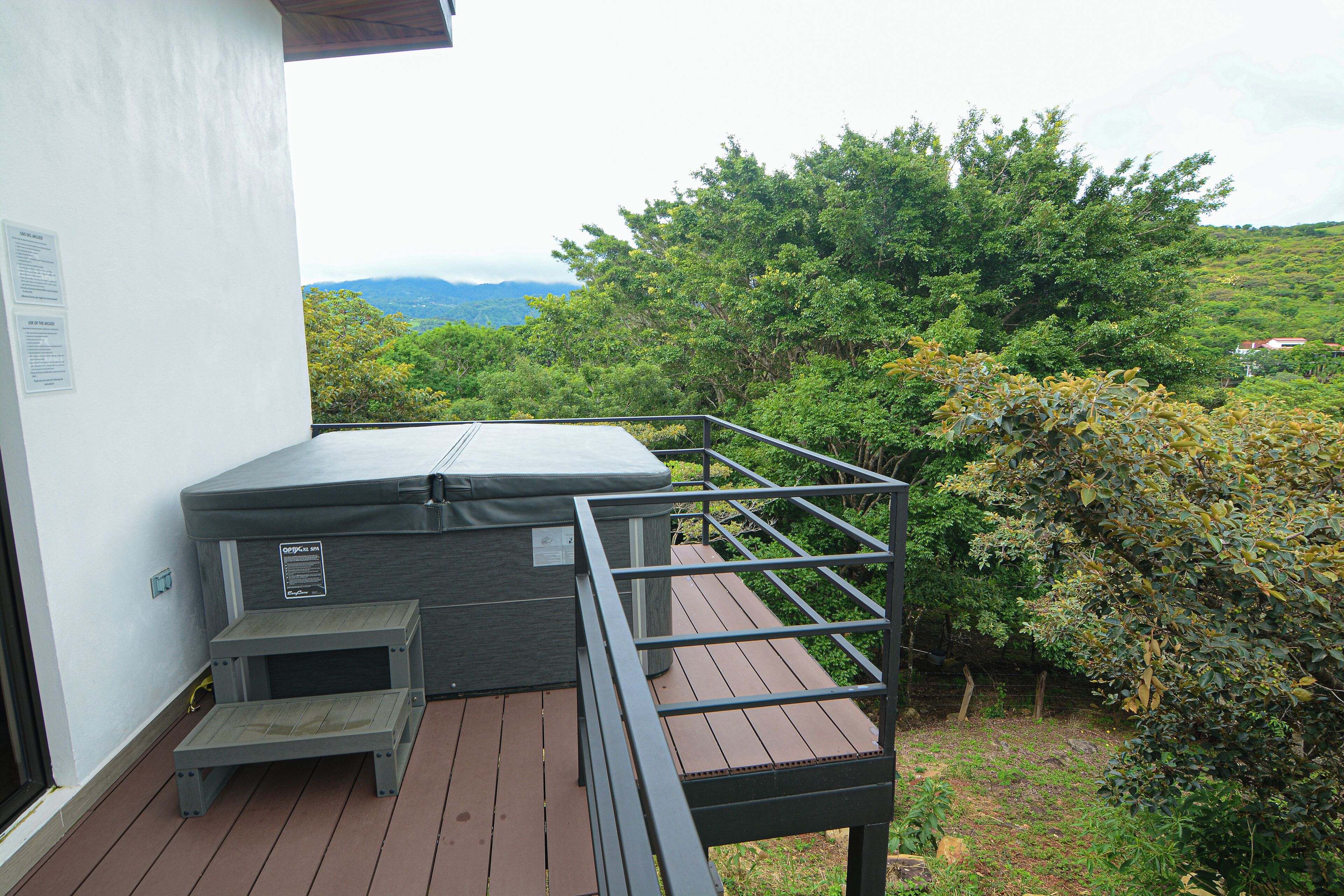Een hottub op een houten terras met een metalen reling, omringd door weelderige begroeiing en een bergachtige achtergrond.