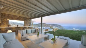 A modern outdoor living area with a stone wall, white furniture, and a view of the sea. - Coastal Retreat With Wellness (Mykonos)