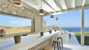 A modern bar area with a stone wall, large windows, and a view of the sea. - Coastal Retreat With Wellness (Mykonos)