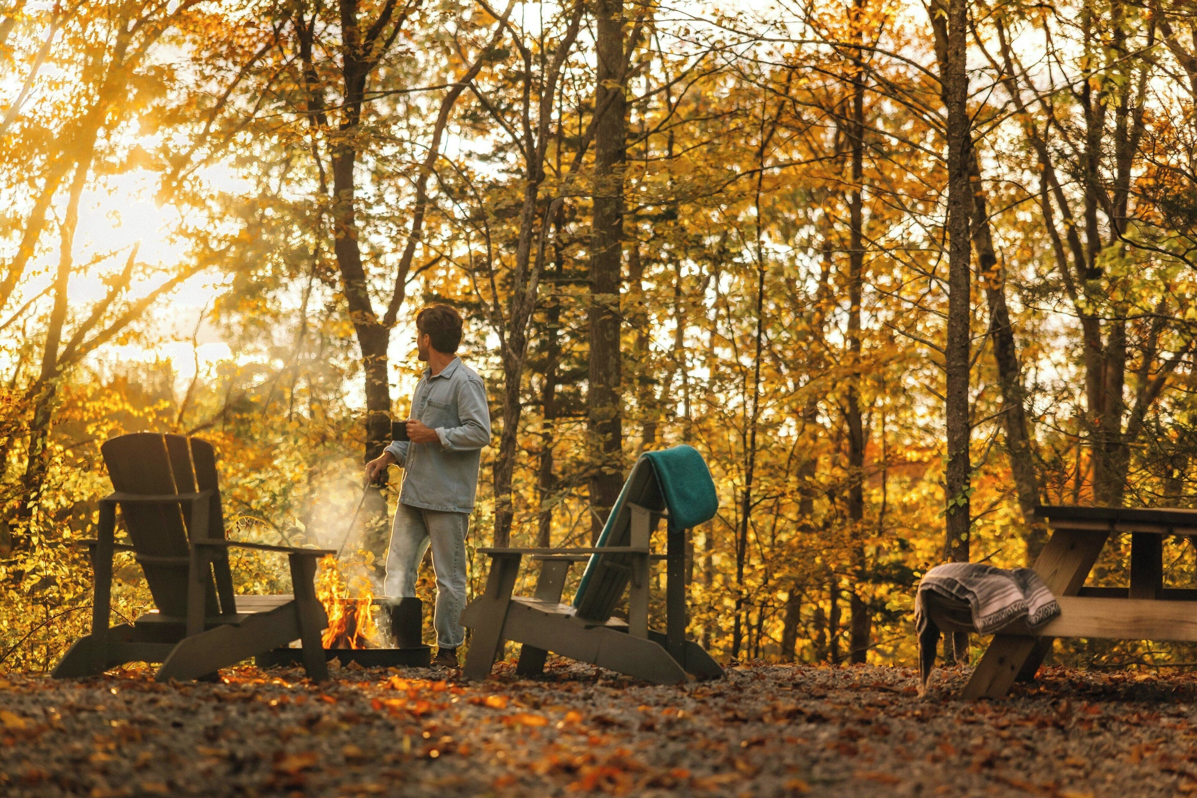 En person står vid ett lägerbål i en skog, bredvid två trästolar och ett picknickbord.