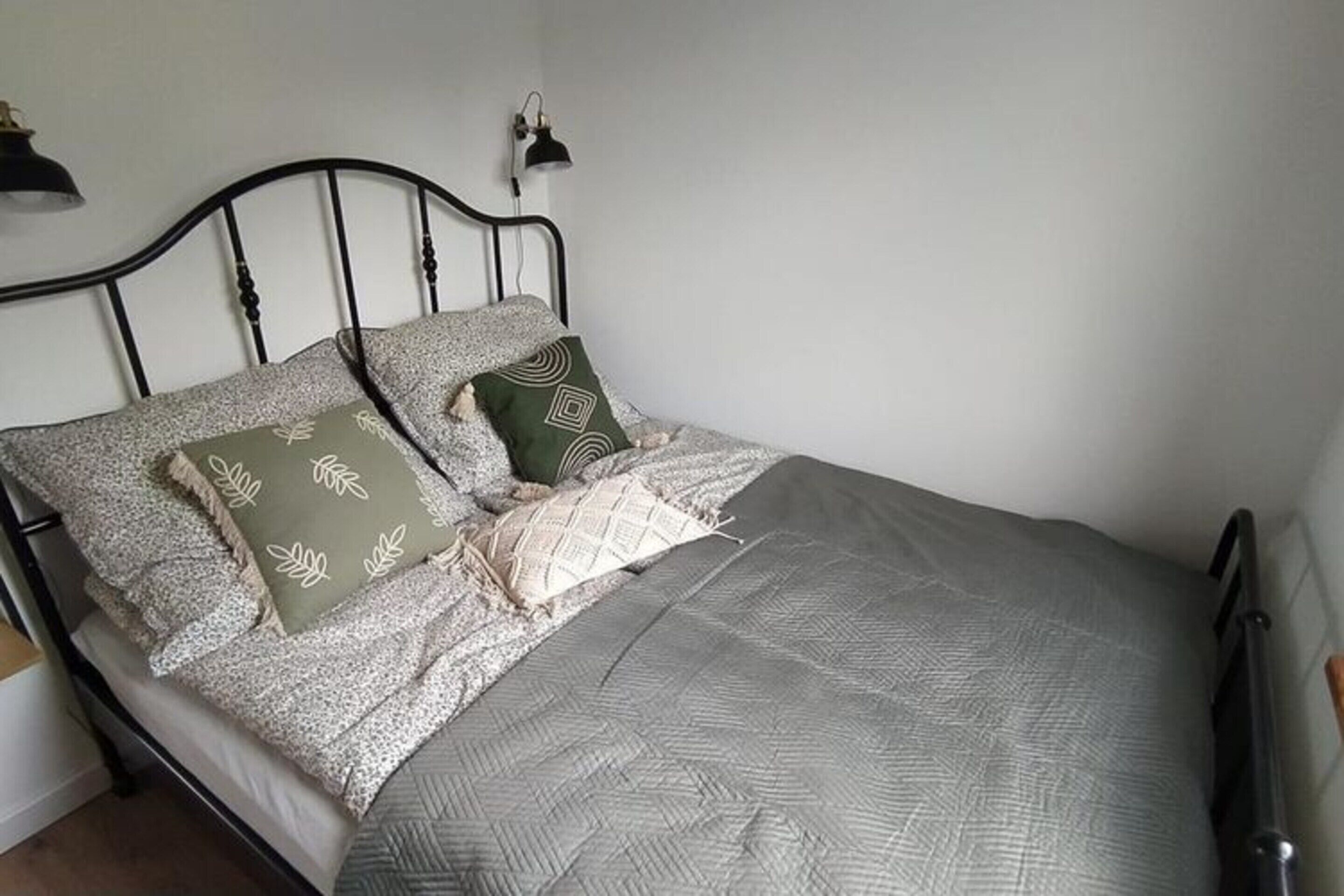A neatly made bed with a grey quilt and patterned pillows, a black metal headboard, and a small bedside lamp.