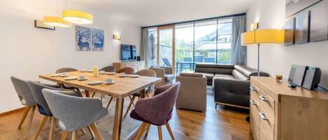 A modern living room with a wooden dining table, grey and purple chairs, a sofa, and a TV on the wall.
