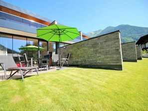 Ein moderner Außenbereich mit Sitzgelegenheit, einer Steinsäule, einem grünen Sonnenschirm und Blick auf Berge.