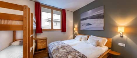 A bedroom with a bunk bed, a single bed, a nightstand, a lamp, a painting, and a window with curtains.